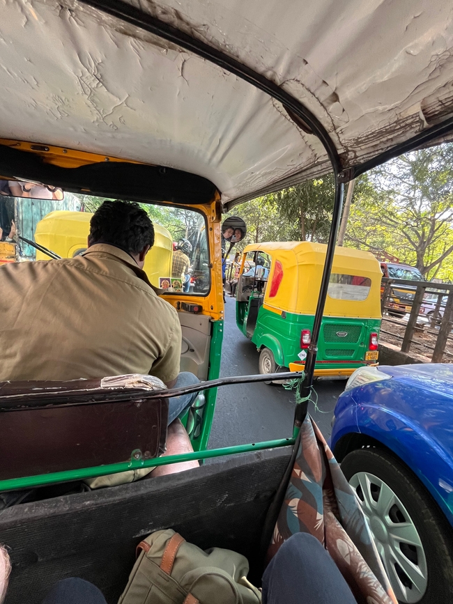 A first-person perspective from inside an auto-rickshaw, commonly known as a tuk-tuk, with a view of the bustling streets featuring other tuk-tuks and vehicles in various colors. The interior shows the back of the driver in a khaki shirt and the edge of the passenger seat, capturing the everyday commute and lively street scene of India.