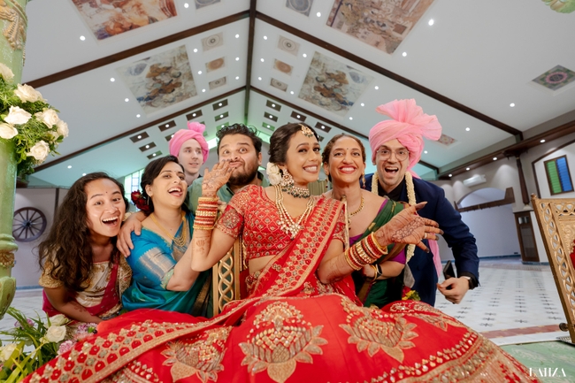 A group of people in vibrant Indian wedding attire are joyfully posing together, with the bride in the center wearing a rich red lehenga, adorned with elaborate jewelry and henna. Our expressions of excitement and the festive turbans highlight the celebratory atmosphere of the occasion.