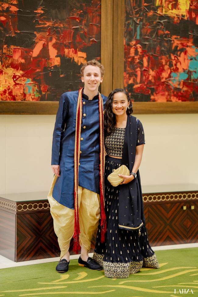 Alex and I are standing next to each other, all smiles. We're dressed in traditional Indian attire, with Alex in a dark blue sherwani and me in a dark blue lehenga with golden detailing.