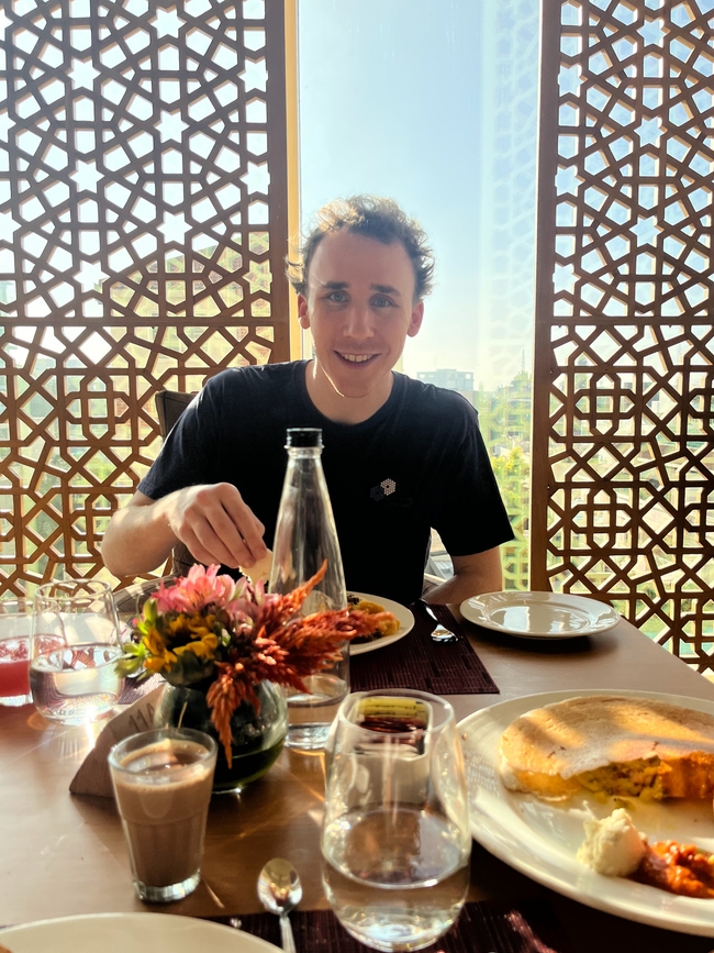 Alex sits at a breakfast table in a sunlit room, smiling at the camera. He's dressed in a casual dark purple T-shirt, and before him on the table is a selection of breakfast items including a glass of water, a hot beverage, and a plate with Indian cuisine. The background features an ornate lattice window that contributes to the bright and airy atmosphere of the setting.