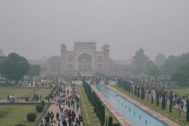 A foggy day at the Taj Mahal, with the grand entrance gate framing a hazy silhouette of this world-famous mausoleum. Visitors are seen milling about, tiny figures against the majestic backdrop, capturing the monument's enduring allure even through the mist.