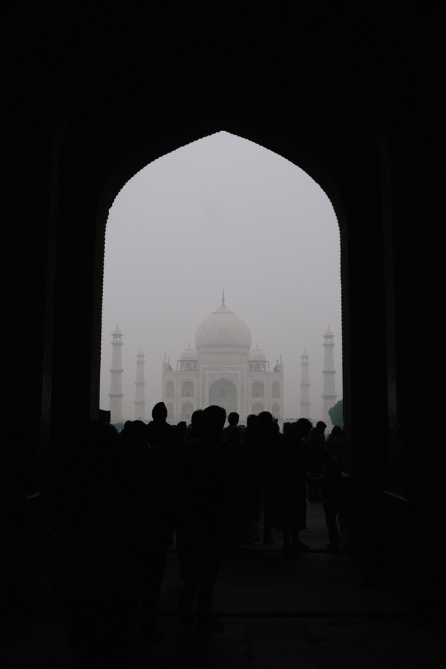 The silhouette of a crowd frames the ethereal form of the Taj Mahal, seen through a grand archway. The iconic mausoleum stands at a distance, shrouded in a light mist, giving the scene a dreamlike quality.